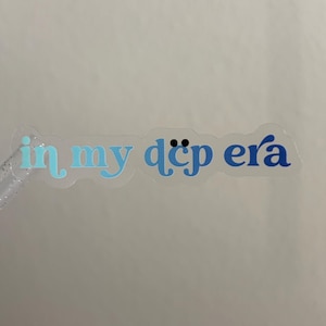 May include: A clear adhesive sticker featuring the text "in my dcp era" in a gradient of blue tones. The letters transition from a light cyan on the left to a deep navy blue on the right. The word "dcp" includes two small black dots above the letter c.