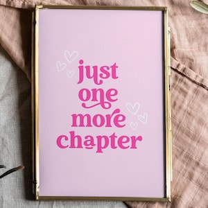 May include: A framed print with a pink background and the text "just one more chapter" in pink lettering. The print is surrounded by a gold frame and is sitting on a light gray surface with a candle and a wooden coaster.