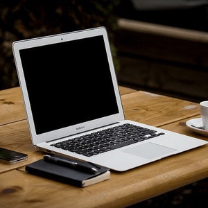May include: A silver laptop with a black screen, a smartphone, a small notebook, a pen, and a cup of coffee on a wooden table. The laptop is open, and the keyboard is visible. The coffee cup is white with a saucer.