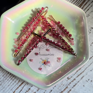 May include: Three gold hair clips with burgundy coloured beads. The clips are on a white and iridescent hexagonal dish. The dish has a sticker with the text "Ly C. Bean Accessories".