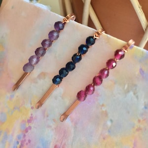 May include: Three hair clips with copper wire and faceted glass beads. The clips are in shades of purple, blue, and pink.