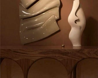 May include: Abstract wall art and a decorative vase. The wall art is a large, cream-coloured, glossy sculpture with a folded, flowing design. A white vase with a curved shape and a cutout detail stands on a wooden console.