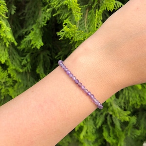 May include: A delicate bracelet made of small, faceted amethyst beads. The bracelet is shown on a person's wrist, with a green leafy background.