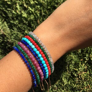 A set of eight beaded bracelets in various colors including turquoise, red, green, blue, and white. The bracelets are made of small, faceted beads and are tied with thin, brown cords.