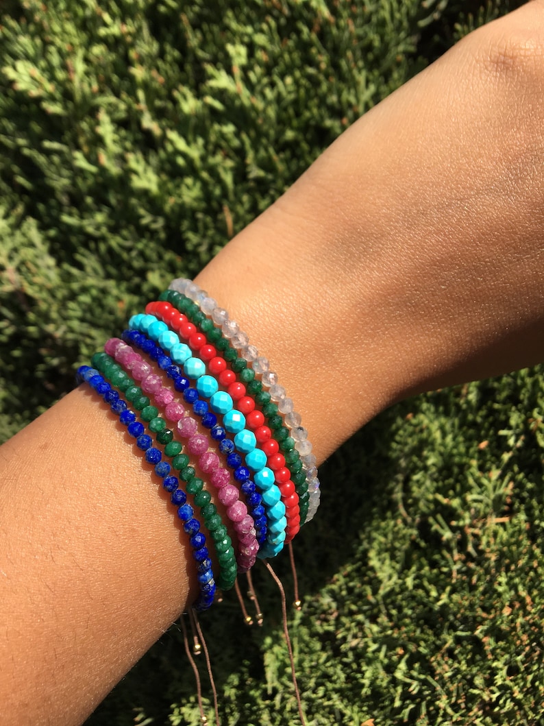 A set of eight beaded bracelets in various colors including turquoise, red, green, blue, and white. The bracelets are made of small, faceted beads and are tied with thin, brown cords.