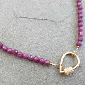 A necklace featuring faceted rubu beads and a gold carabiner clasp. The clasp is teardrop-shaped with small, clear stones. The necklace is displayed on a gray textured surface.  The purple gemstone beads are uniform in size and shape, creating a consistent look. The gold clasp adds a touch of elegance to the overall design. This stylish necklace is perfect for adding a touch of color and sophistication to any outfit.