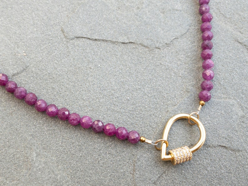 A necklace featuring faceted rubu beads and a gold carabiner clasp. The clasp is teardrop-shaped with small, clear stones. The necklace is displayed on a gray textured surface.  The purple gemstone beads are uniform in size and shape, creating a consistent look. The gold clasp adds a touch of elegance to the overall design. This stylish necklace is perfect for adding a touch of color and sophistication to any outfit.