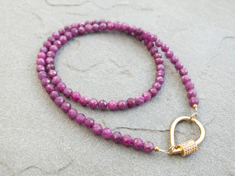Faceted ruby gemstone necklace with a teardrop gold carabiner clasp. The necklace is made of 4mm small, faceted beads and has a simple, elegant design. The gold clasp is a unique feature that adds to the overall look of the necklace. This purple gemstone necklace is perfect for adding a touch of elegance to any outfit.