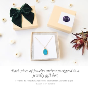 A jewelry displayed inside a kraft jewelry gift box with a white insert, surrounded by dried flowers and two additional branded boxes—one topped with a dark green velvet bow. The background is white, styled for an elegant and minimal gift presentation.