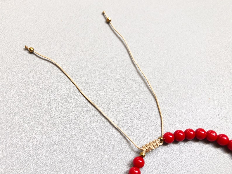 A red beaded bracelet with a sliding knot closure. The bracelet is made of small, round 4mm coral beads and is strung on a thin, beige cord.