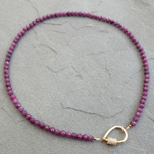 A necklace made of faceted, dark purplish-pink ruby gemstone beads. The necklace has a gold tear-drop shape carabiner clasp. The beads are small and uniform in size. The necklace is displayed on a gray textured surface.