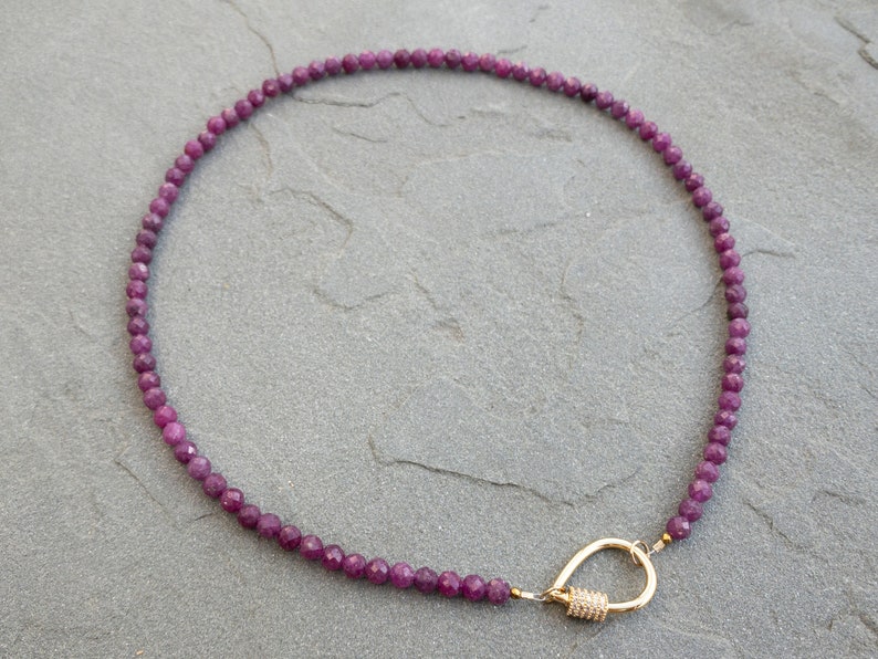 A necklace made of faceted, dark purplish-pink ruby gemstone beads. The necklace has a gold tear-drop shape carabiner clasp. The beads are small and uniform in size. The necklace is displayed on a gray textured surface.