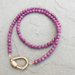 A necklace featuring 4mm faceted, dark pink gemstone beads. The necklace has a gold carabiner clasp with small, clear cubic zirconia accents. The necklace is displayed on a gray textured surface.