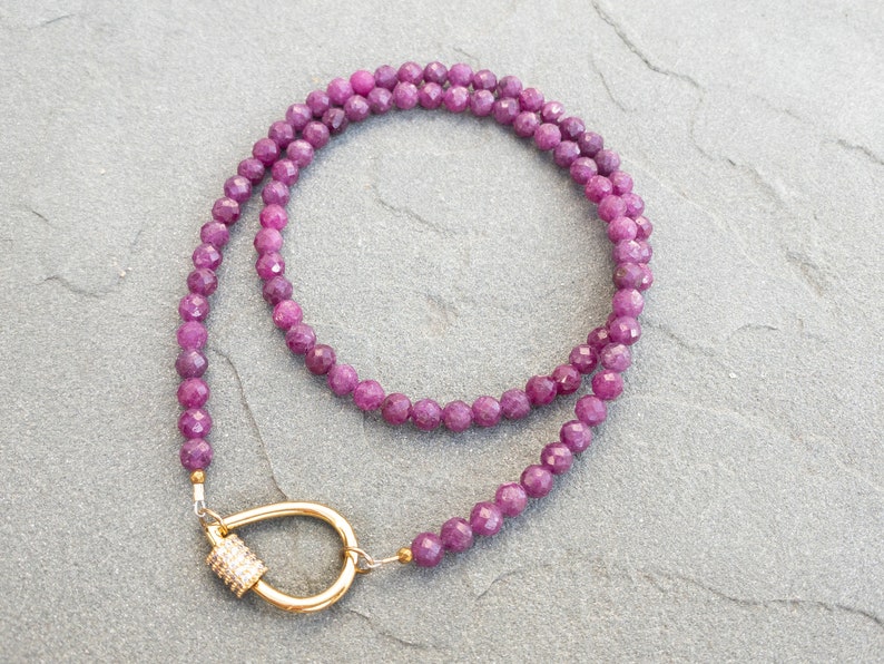A necklace featuring 4mm faceted, dark pink gemstone beads. The necklace has a gold carabiner clasp with small, clear cubic zirconia accents. The necklace is displayed on a gray textured surface.