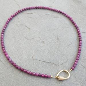 A necklace made of faceted, dark pink ruby gemstone beads and a gold clasp. The necklace is displayed on a gray textured surface. The beads are 4mm and uniform in size and shape, creating a delicate and elegant look. The gold clasp is a simple carabiner, yet stylish design that complements the gemstone beads. The necklace is approximately 15 inches in length, making it a versatile accessory for everyday wear or special occasions.
