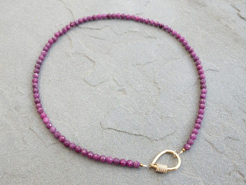 A necklace made of faceted, dark pink ruby gemstone beads and a gold clasp. The necklace is displayed on a gray textured surface. The beads are 4mm and uniform in size and shape, creating a delicate and elegant look. The gold clasp is a simple carabiner, yet stylish design that complements the gemstone beads. The necklace is approximately 15 inches in length, making it a versatile accessory for everyday wear or special occasions.