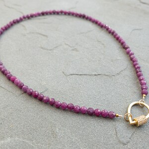 A necklace featuring faceted, dark pink ruby gemstone beads and a gold clasp. The necklace is displayed on a gray textured surface. The beads are uniformly sized and create a continuous strand. The gold clasp is a carabiner design with a texture dtear-drop shaped accent.