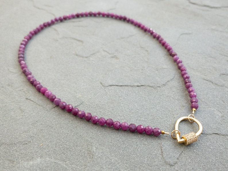 A necklace featuring faceted, dark pink ruby gemstone beads and a gold clasp. The necklace is displayed on a gray textured surface. The beads are uniformly sized and create a continuous strand. The gold clasp is a carabiner design with a texture dtear-drop shaped accent.