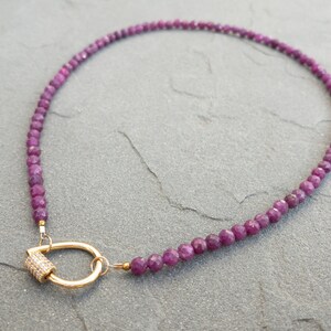 A necklace made of 4mm faceted, dark pink gemstone beads and a gold carabiner charm holder clasp. The clasp is a gold-toned tear-drop shaped with a small, pave-set diamond cylinder. The necklace is displayed on a gray textured surface.