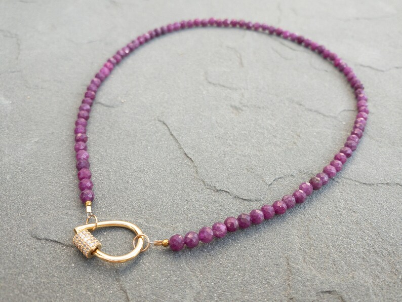 A necklace made of 4mm faceted, dark pink gemstone beads and a gold carabiner charm holder clasp. The clasp is a gold-toned tear-drop shaped with a small, pave-set diamond cylinder. The necklace is displayed on a gray textured surface.