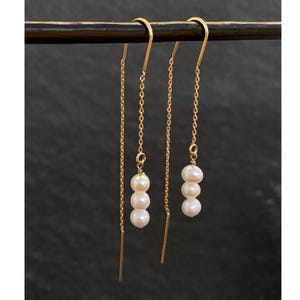 May include: Gold-toned threader earrings with three white pearls suspended from a delicate chain. The earrings are displayed against a dark background, showcasing their elegant design and simplicity. The earrings are a stylish accessory.