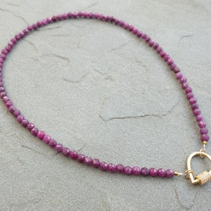 A necklace featuring faceted, dark pink ruby gemstone beads and a gold clasp. The necklace is displayed on a gray textured surface. The clasp is a gold-toned, tear-drop design with small, clear accents. The necklace is approximately 16 inches in length, suitable for everyday wear or special occasions. The gemstone beads are uniform in size and shape, creating an elegant look. The overall style is minimalist and sophisticated, perfect for adding a touch of color and sparkle to any outfit.