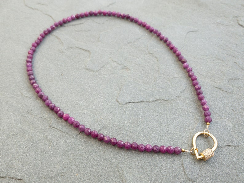 A necklace featuring faceted, dark pink ruby gemstone beads and a gold clasp. The necklace is displayed on a gray textured surface. The clasp is a gold-toned, tear-drop design with small, clear accents. The necklace is approximately 16 inches in length, suitable for everyday wear or special occasions. The gemstone beads are uniform in size and shape, creating an elegant look. The overall style is minimalist and sophisticated, perfect for adding a touch of color and sparkle to any outfit.