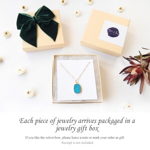 A jewelry displayed inside a kraft jewelry gift box with a white insert, surrounded by dried flowers and two additional branded boxes, one topped with a dark green velvet bow. The background is white, styled for an elegant and minimal gift presentation.