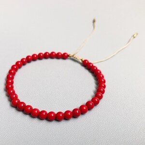 A red beaded bracelet with a sliding knot closure. The bracelet is made of small, round 4mm coral beads and is strung on a thin, beige cord.