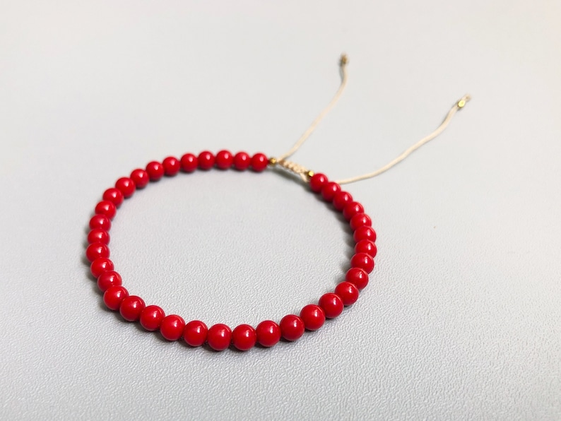 A red beaded bracelet with a sliding knot closure. The bracelet is made of small, round 4mm coral beads and is strung on a thin, beige cord.