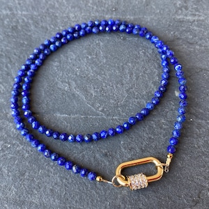 Può includere: Una collana di perline di lapislazzuli blu con una chiusura dorata. La chiusura è una chiusura a moschettone grande, ovale, con strass trasparenti.