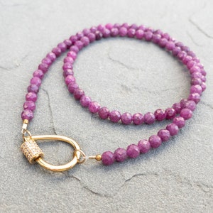 A delicate necklace featuring faceted, dark pink ruby gemstone beads and a gold clasp. The necklace is elegantly draped on a textured gray surface. The clasp is a gold-toned, tear-drop shaped, curved bar with a small, sparkly cylinder accent. The overall aesthetic is minimalist and sophisticated, suitable for everyday wear or special occasions. The necklace's length is approximately 16 inches, ideal for layering or wearing alone.