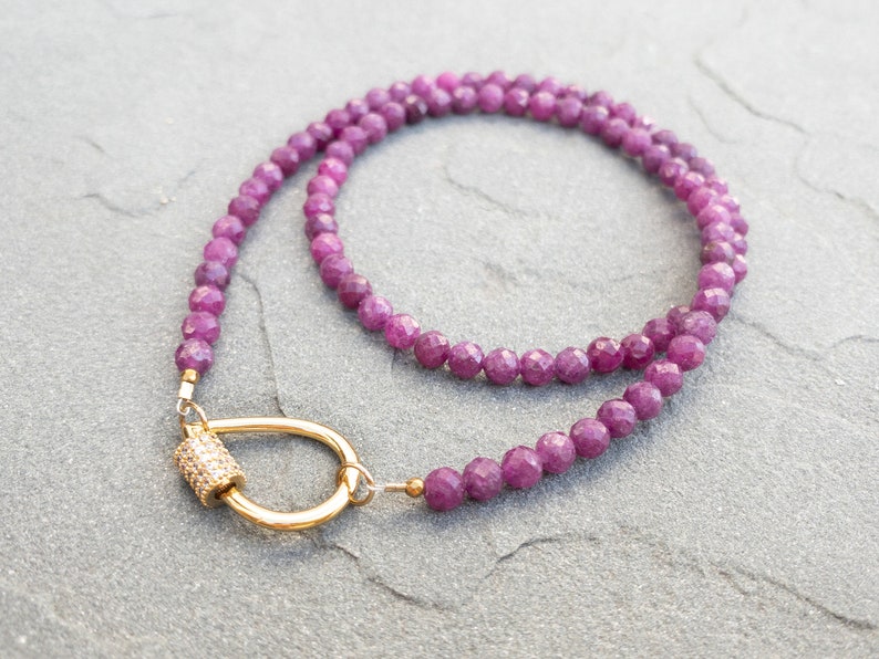A delicate necklace featuring faceted, dark pink ruby gemstone beads and a gold clasp. The necklace is elegantly draped on a textured gray surface. The clasp is a gold-toned, tear-drop shaped, curved bar with a small, sparkly cylinder accent. The overall aesthetic is minimalist and sophisticated, suitable for everyday wear or special occasions. The necklace's length is approximately 16 inches, ideal for layering or wearing alone.