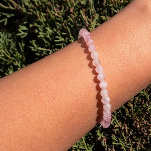 May include: A delicate bracelet made of pale pink and white gemstone beads. The bracelet is worn on a person's wrist.