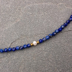 Lapis lazuli beaded necklace details. There are 3mm faceted navy blue lapis lazuli beads and a white pearl in the middle. There are 2mm gold hematite beads at each side of the pearl.