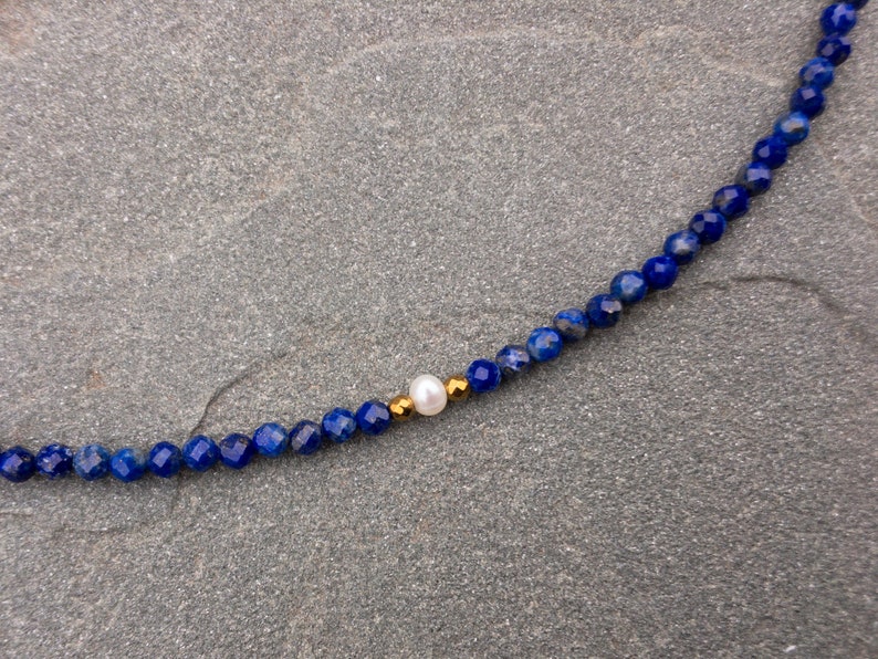 Lapis lazuli beaded necklace details. There are 3mm faceted navy blue lapis lazuli beads and a white pearl in the middle. There are 2mm gold hematite beads at each side of the pearl.