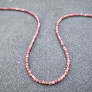 Può includere: Una collana di pietre preziose rosa realizzata con piccole perle rotonde. La collana è su uno sfondo grigio.