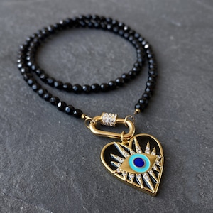 Peut inclure: Un collier de perles d'onyx noir avec un pendentif en forme de cœur doré orné d'un œil maléfique. Le pendentif a un œil en émail bleu et turquoise avec des strass clairs tout autour.