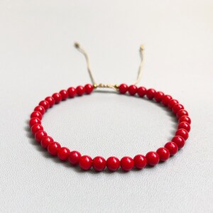 A red beaded bracelet with a sliding knot closure. The bracelet is made of small, round 4mm coral beads and is strung on a thin, beige cord.