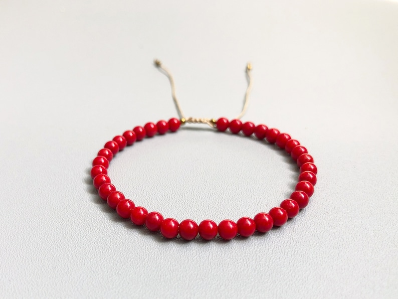 A red beaded bracelet with a sliding knot closure. The bracelet is made of small, round 4mm coral beads and is strung on a thin, beige cord.