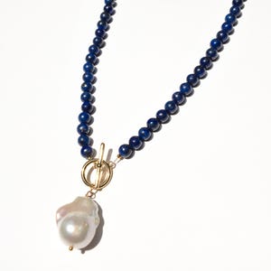 May include: A necklace featuring a strand of small, dark blue beads. The necklace has a gold-colored clasp and a large, irregularly shaped white pearl pendant. The jewelry is displayed against a white background.