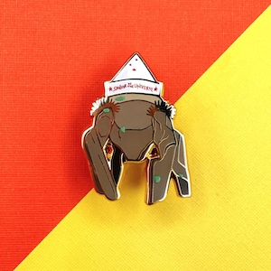 May include: A metal enamel pin featuring a brown, four-legged creature with green spots, wearing a white pointed hat with red stars and the text Sailor of the Universe in red lettering. The pin has gold-toned metal outlines and edges.