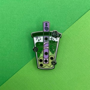 Puede incluir: Un pin de esmalte verde, morado y blanco de una taza de boba tea con perlas de tapioca, una pajita y una flor en la parte superior. El pin tiene un acabado plateado brillante.