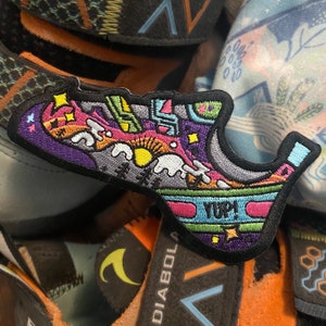 Puede incluir: Un parche bordado colorido que representa un zapato con una escena de montaña y la palabra "YUP!" en una pancarta. El parche es negro con una variedad de colores, incluyendo rosa, verde, amarillo y azul.