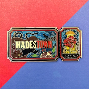 Puede incluir: Un pin de esmalte rectangular con forma de boleto con el texto HADESTOWN. El diseño presenta una guitarra marrón, un pájaro negro, una granada y una flor roja bajo una cúpula amarilla. El lado derecho incluye el texto $20.06 en letras doradas.