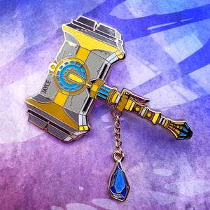 May include: An enamel pin depicting a hammer in grey, yellow, and blue. The pin has the name "JAYCE" and a dangling blue gem. The hammer design is highly detailed. The pin is suitable for clothing or accessories.