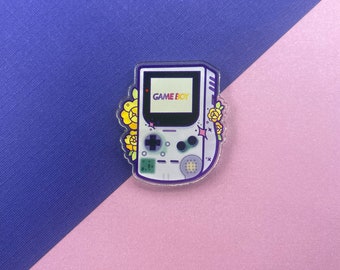May include: A white Game Boy Color with purple accents and yellow flowers. The screen reads "GAME BOY".