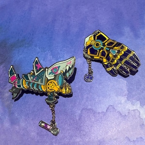 May include: Two enamel pins, one featuring a blue, yellow and pink robotic shark with a chain and a small pink and purple charm, and the other featuring a gold and purple robotic hand with a chain and a small blue charm.