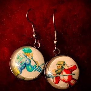 May include: A pair of round earrings with silver-colored hooks. Each earring features a small, circular image of a person playing a wind instrument. One figure is dressed in blue and green, the other in red. The background is a deep red.