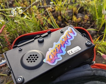 May include: A black box with a speaker and a holographic sticker that says "Deep Party" on it. The box is in a red and black carrying case. The box is sitting on a bed of green grass and rocks.
